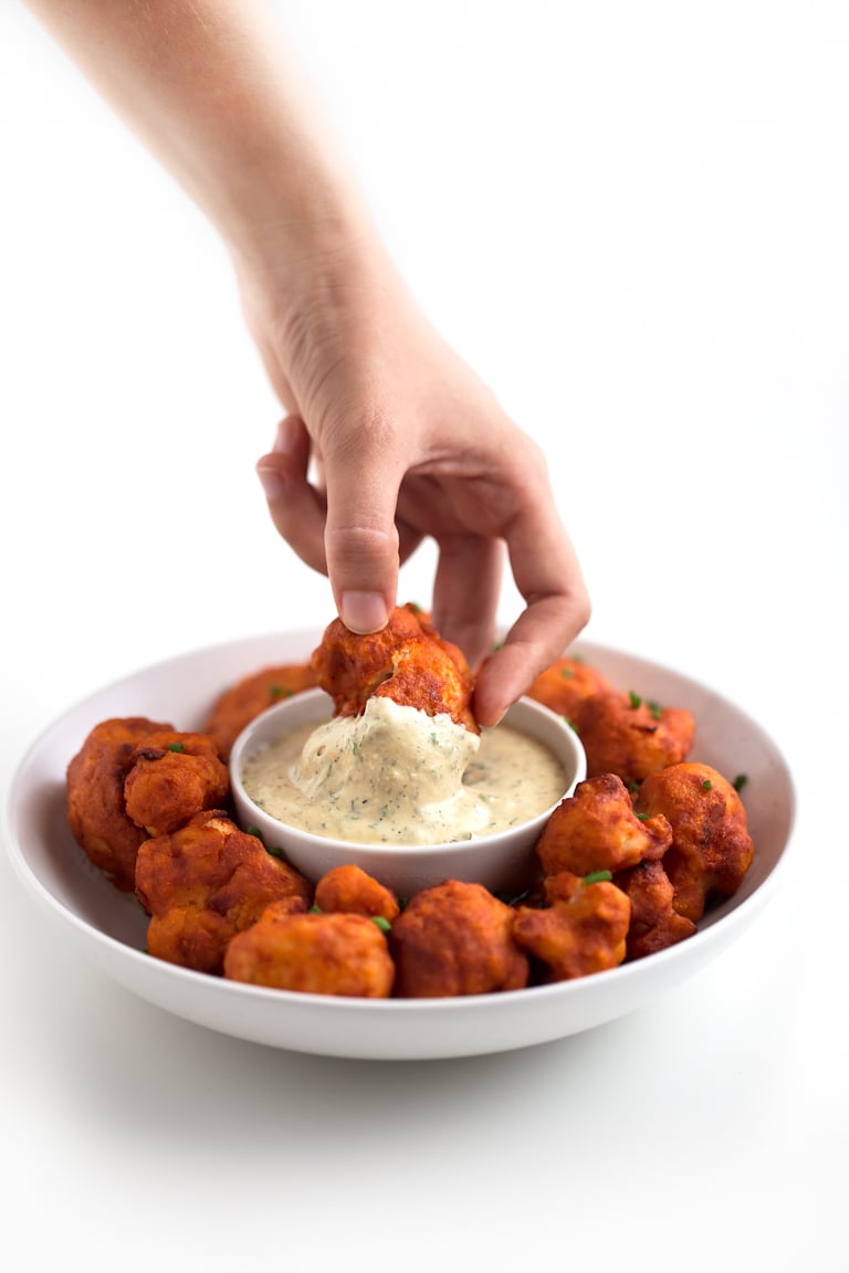 Photo of a bowl of buffalo cauliflower wings being dipped in vegan ranch dressing