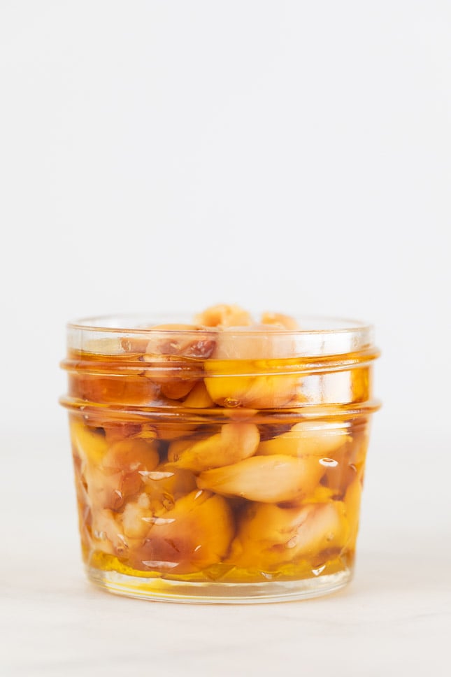 A side shot of a glass jar with roasted garlic cloves and covered with oil
