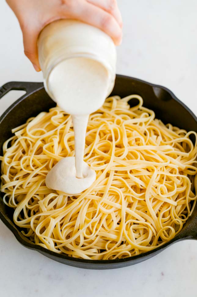 A picture of a hand with a jar pouring some vegan Alfredo sauce onto a skillet with cooked pasta