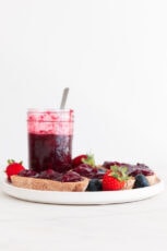 A side shot of a dish with a toast and a jar with berry compote and fresh berries around