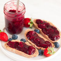 A square picture of a jar and some bread slices with berry compote onto a dish