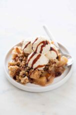 Photo of a dish with a spoon and some vegan apple crisp topped with vegan ice cream and a drizzle of vegan caramel.
