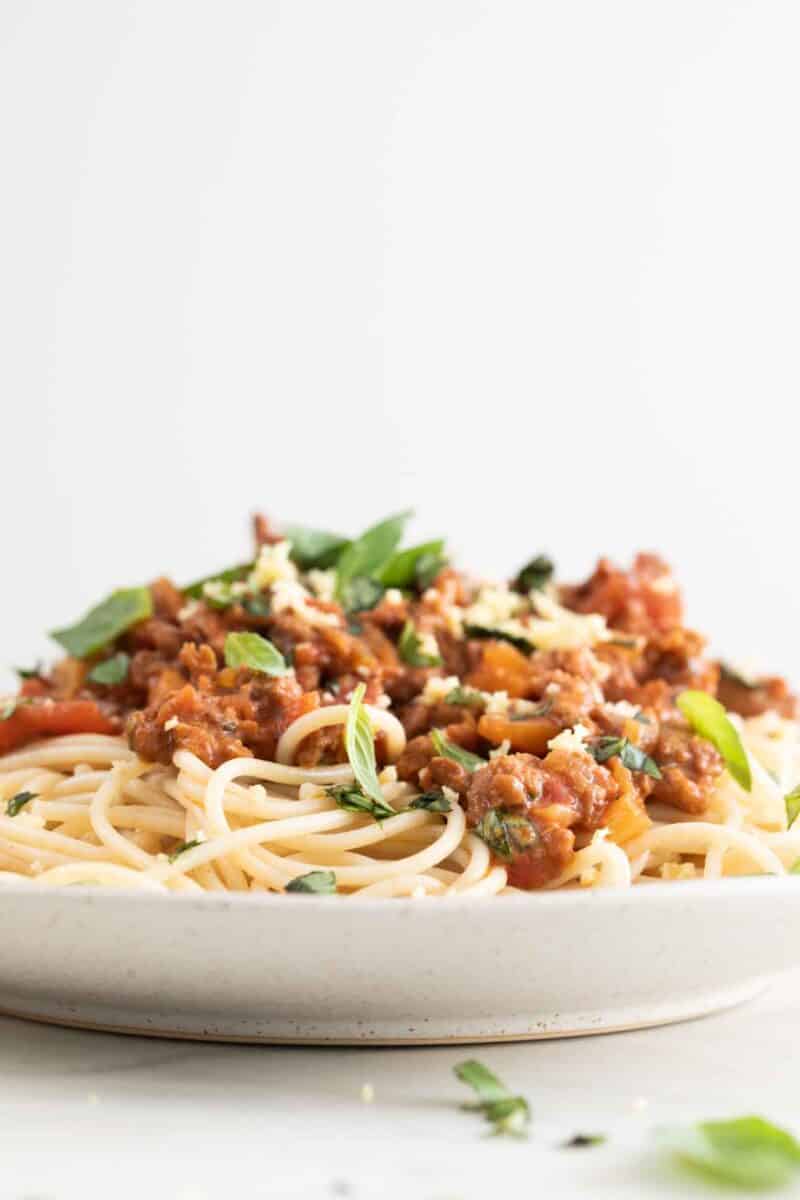 Side picture of a dish with vegan bolognese over cooked spaghetti and garnished with some vegan Parmesan cheese and fresh basil