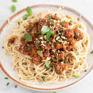 Square photo of a dish with vegan bolognese sauce with spaghetti and with some vegan Parmesan cheese and fresh basil on top