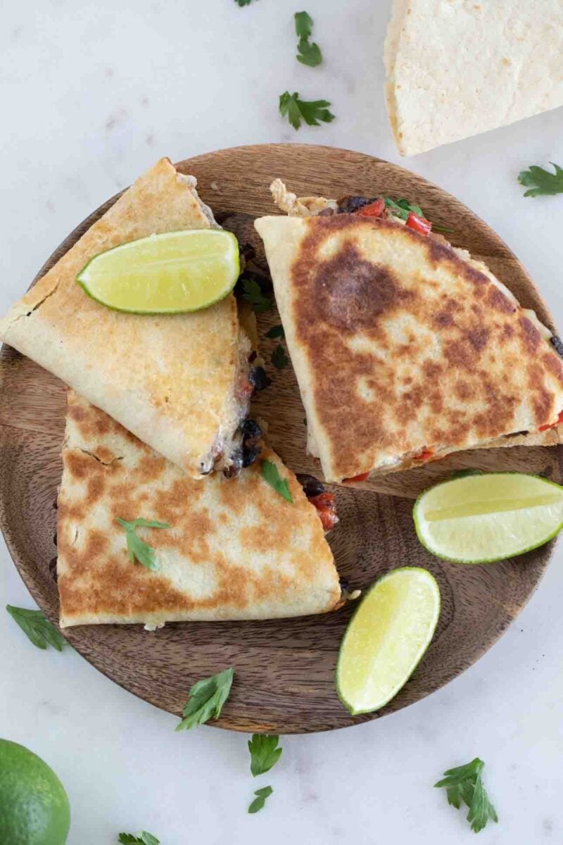 Photo from above of 3 pieces of vegan quesadilla onto a wooden board with some chopped cilantro and lime wedges