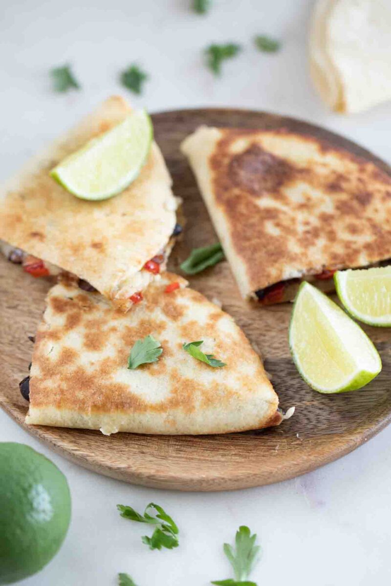 Photo of 3 pieces of vegan quesadilla onto a wooden board with some chopped cilantro and lime wedges
