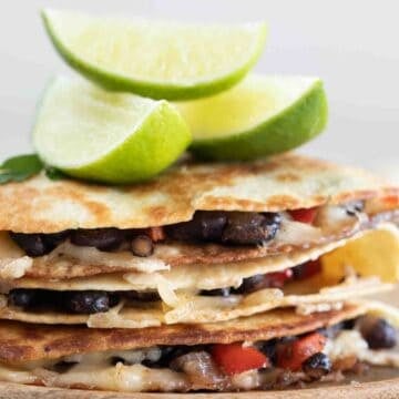 Side photo of a pile of vegan quesadillas onto a wooden board with some lime wedges
