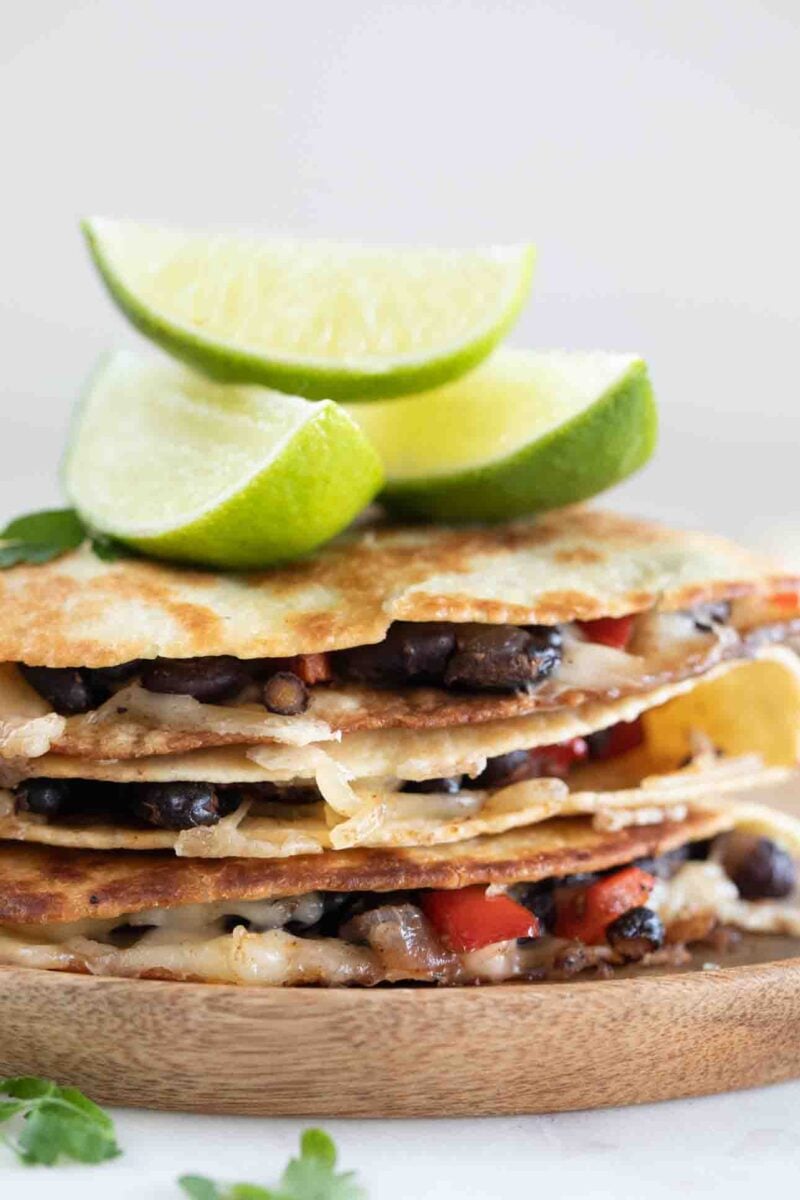 Side photo of a pile of vegan quesadillas onto a wooden board with some lime wedges