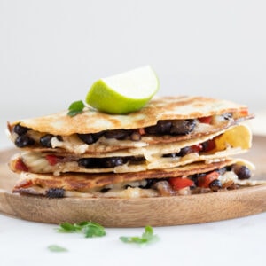 Square photo of a pile of vegan quesadillas onto a wooden board with a lime wedge