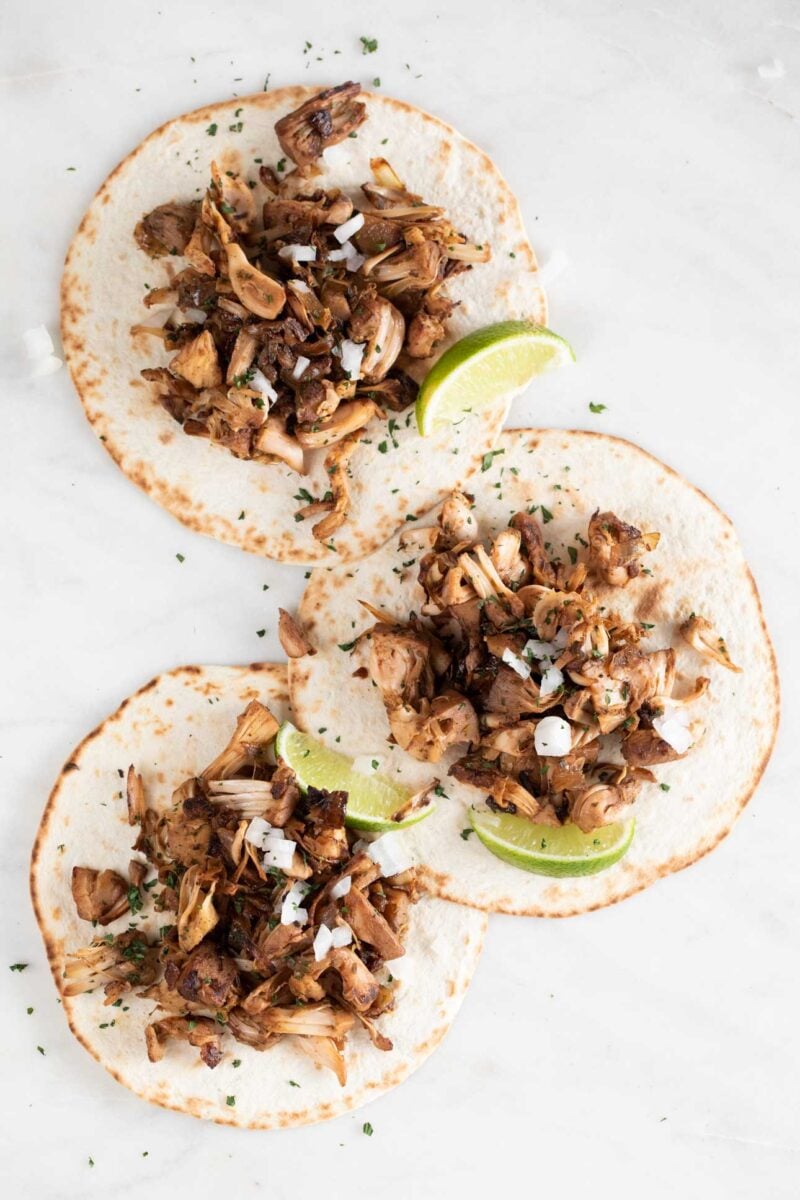 Overview photo of 3 jackfruit carnitas with some lime wedges, chopped cilantro and diced onion