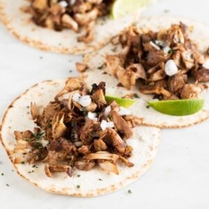 Square photo of 3 jackfruit carnitas served with tortillas, lime wedges, diced onion and chopped cilantro
