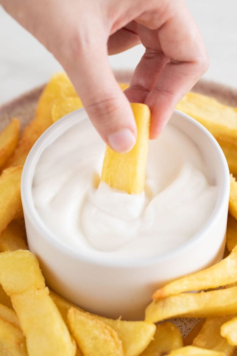 Picture a plate with french fries and a small bowl with vegan aioli and a hand dipping into the sauce.