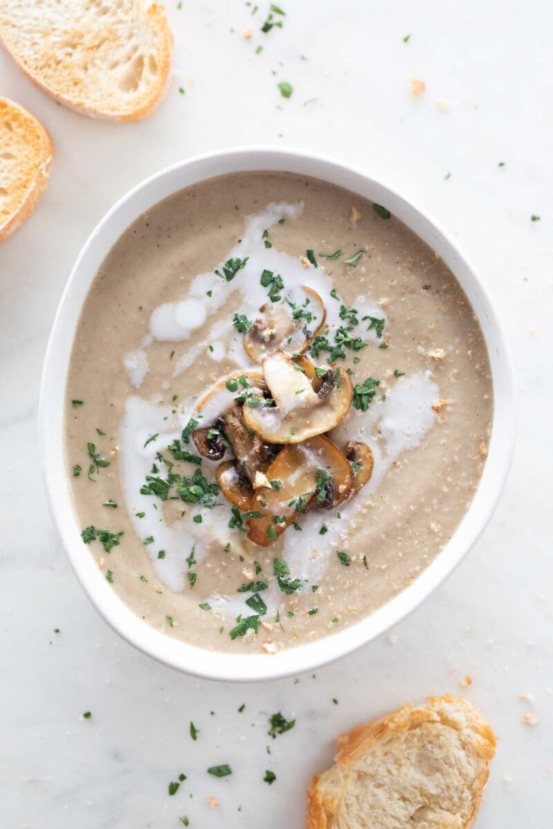 Overview photo of a bowl of vegan mushroom soup with some bread slices, chopped parsely, a drizzle of coconut milk and some sautéed mushrooms.