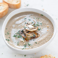 Square photo of a bowl of vegan mushroom soup, topped with some sautéed mushrooms, chopped parsley, and a drizzle of coconut milk.