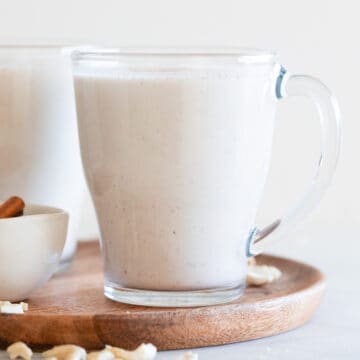 Two mugs of vegan eggnog onto a wooden board with some cashews and cinnamon sticks around.