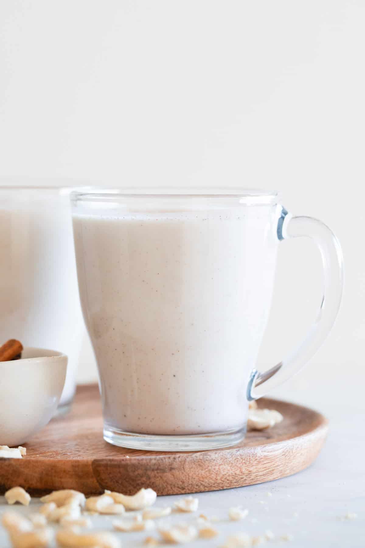 Two mugs of vegan eggnog onto a wooden board with some cashews and cinnamon sticks around.