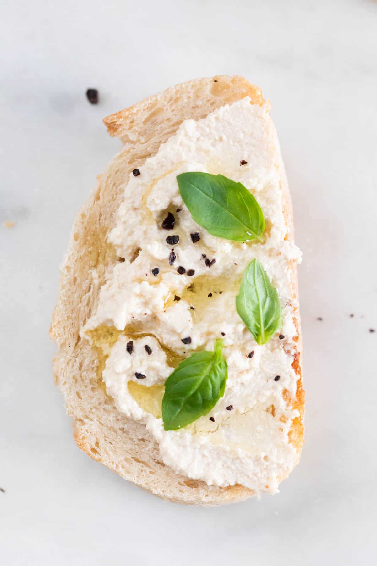 A slice of bread spread with some vegan tofu ricotta cheese and some basil leaves, black pepper and olive oil on top.