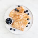 Overview of a plate with several folded vegan crepes topped with fresh blueberries and powdered sugar.