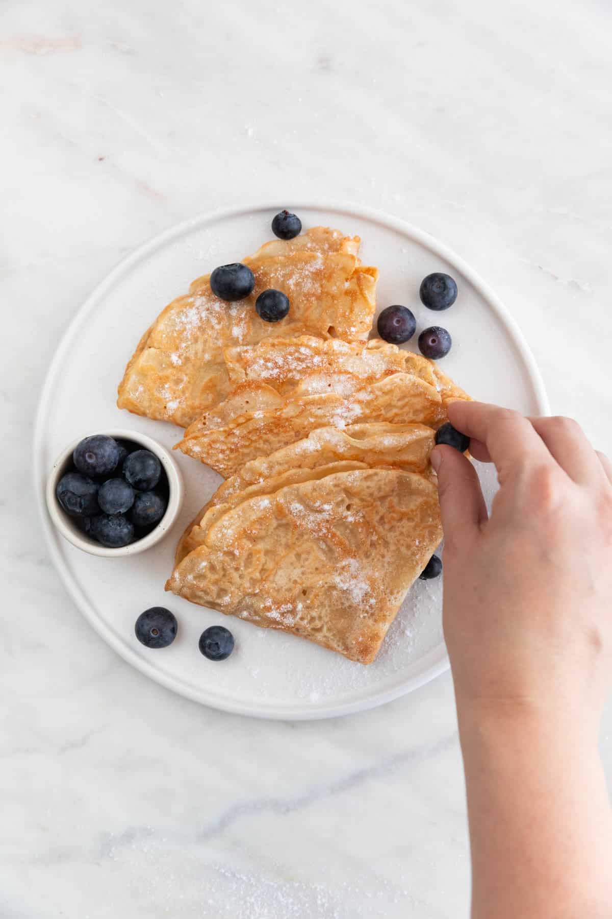 A hand adding fresh blueberries onto a plate with folded vegan crepes topped with powdered sugar.