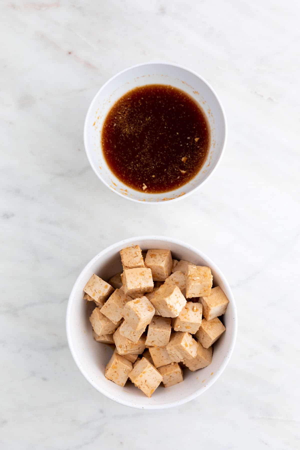 Marinated tofu cubes in a bowl and a bowl with the marinade liquid after draining the tofu cubes.