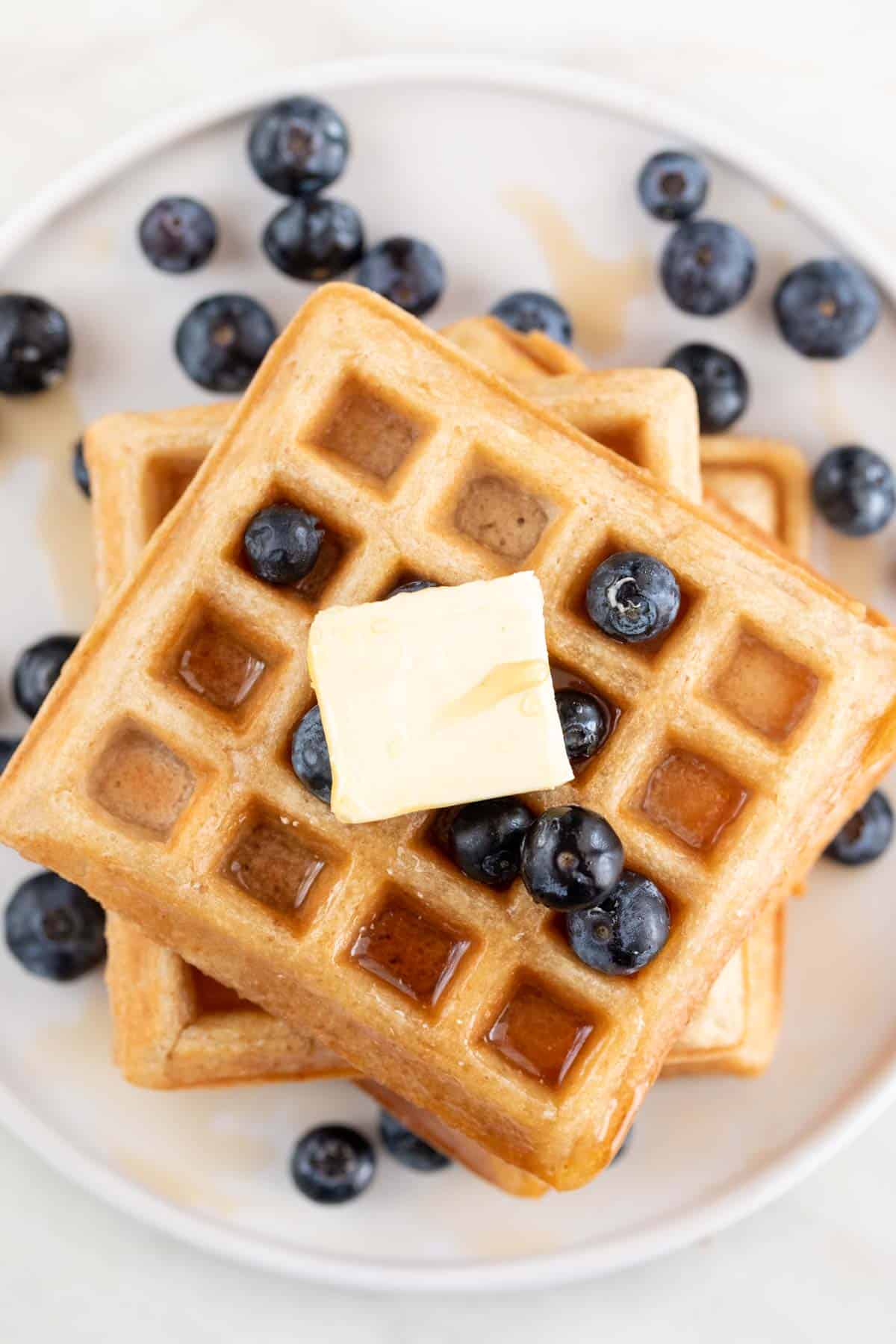 A tower of vegan waffles topped with blueberries, vegan butter, and maple syrup.