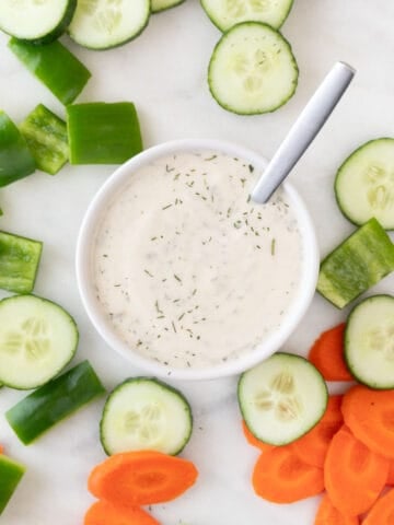 A spoon rests in a bowl of vegan ranch dressing, surrounded by crudités.