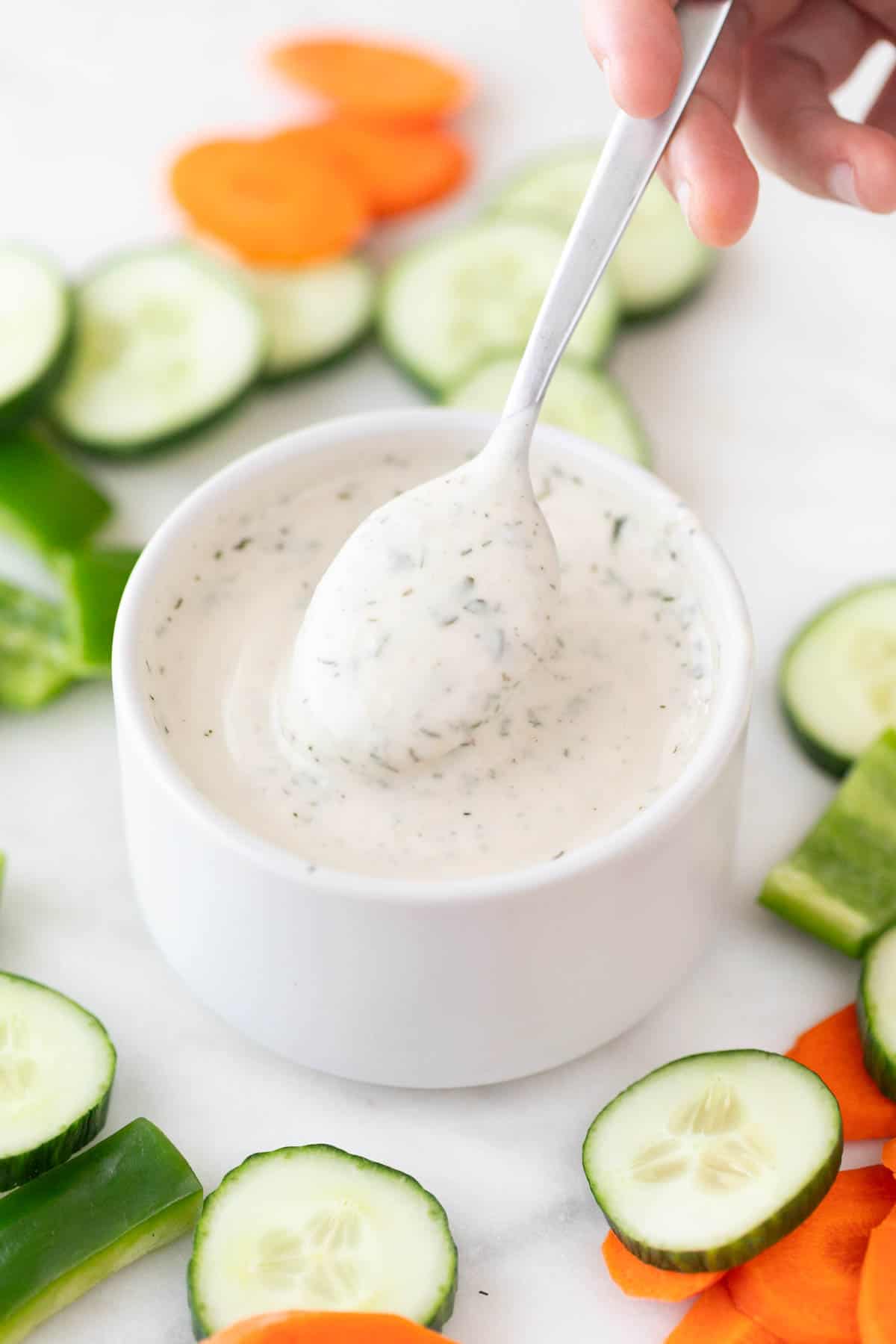A hand dipping a spoon into a bowl of vegan ranch dressing surrounded by crudités.