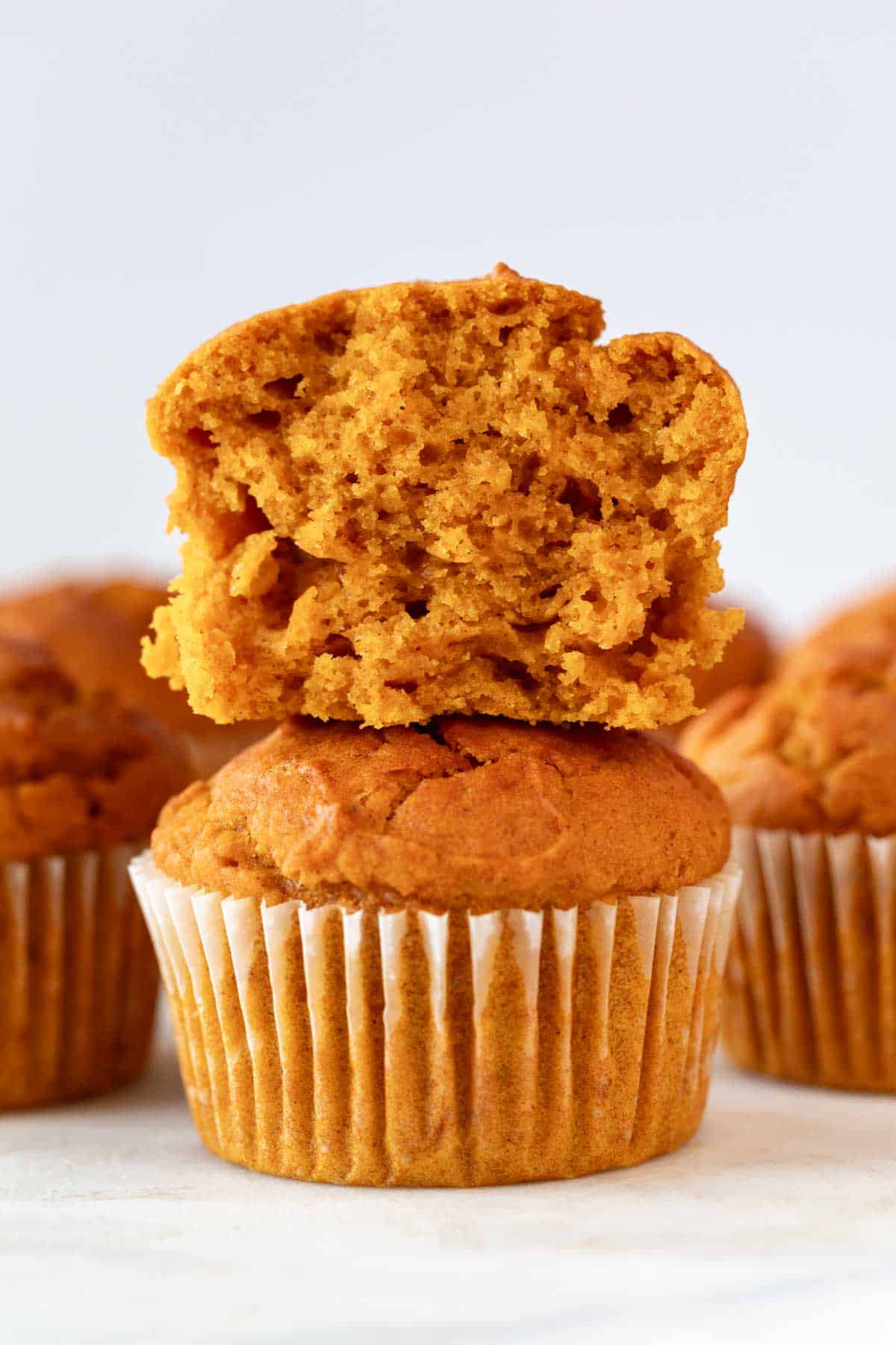 Vegan pumpkin muffins stacked, one sliced atop, surrounded by more muffins.