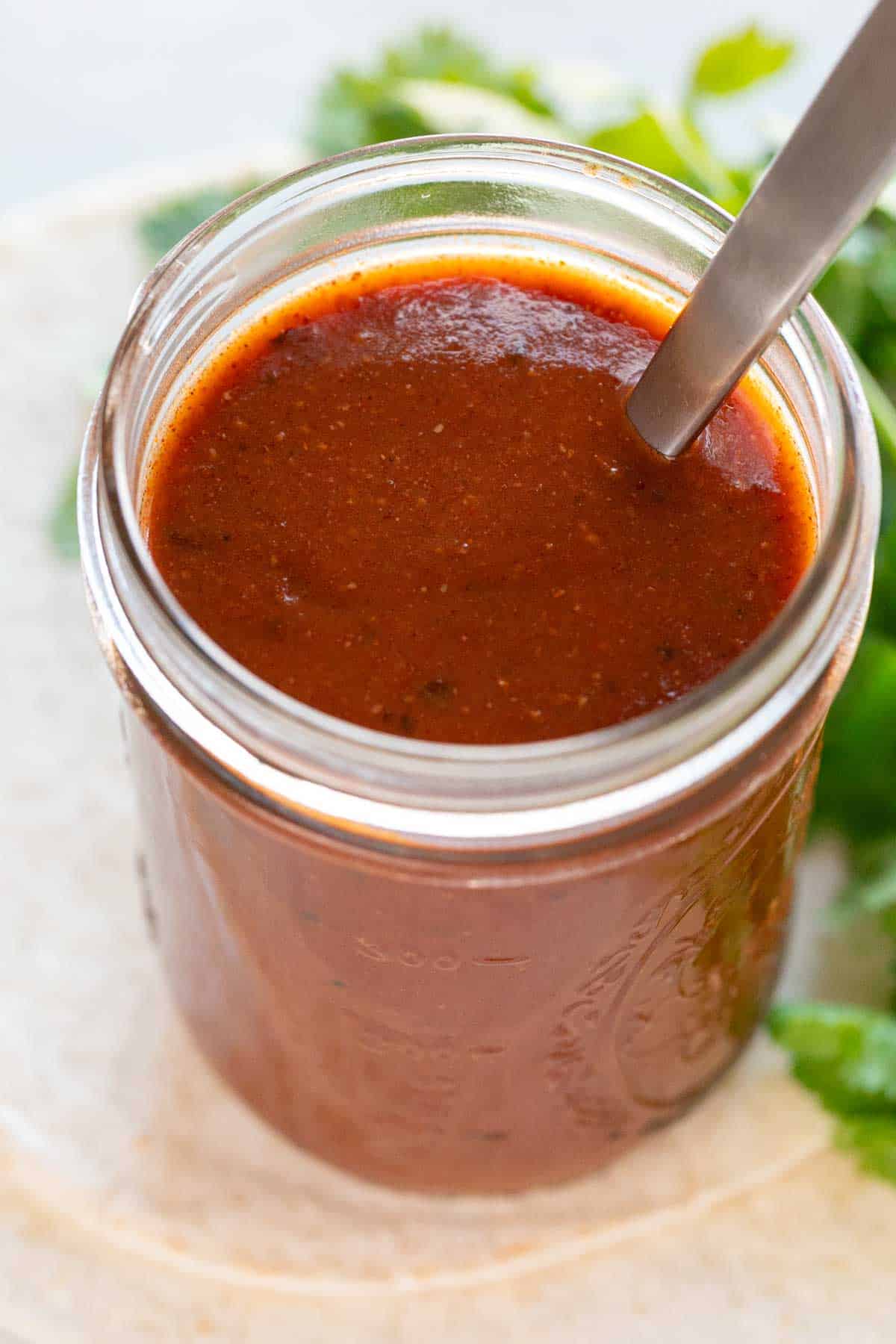 Jar of red enchilada sauce with a spoon, tortillas, and cilantro arranged around it.
