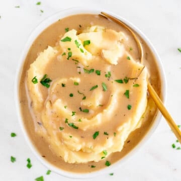 A bowl containing mashed potatoes, vegan gravy, parsley garnish, and a serving spoon.