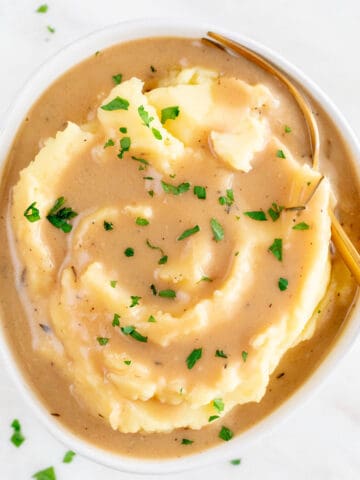 A bowl containing mashed potatoes, vegan gravy, parsley garnish, and a serving spoon.