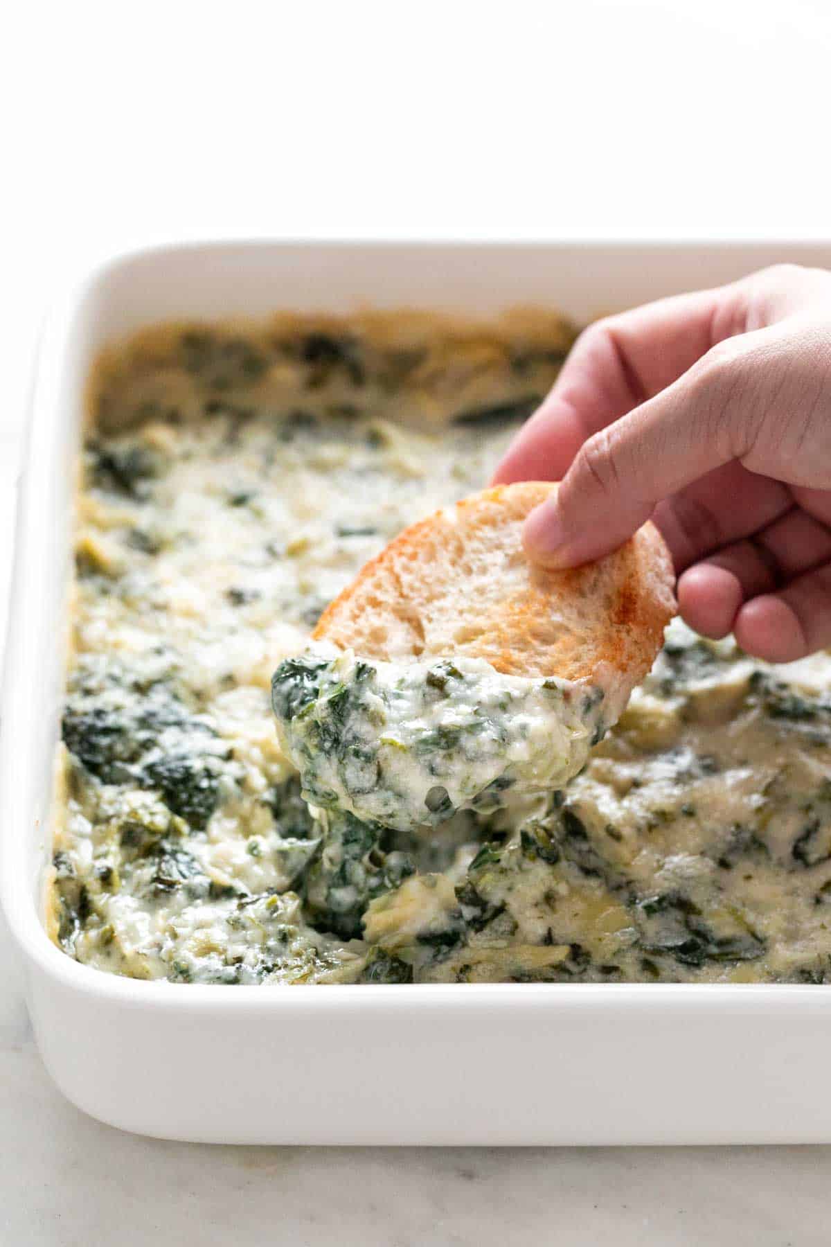 Hand dunking a slice of bread into a dish of vegan spinach artichoke dip.
