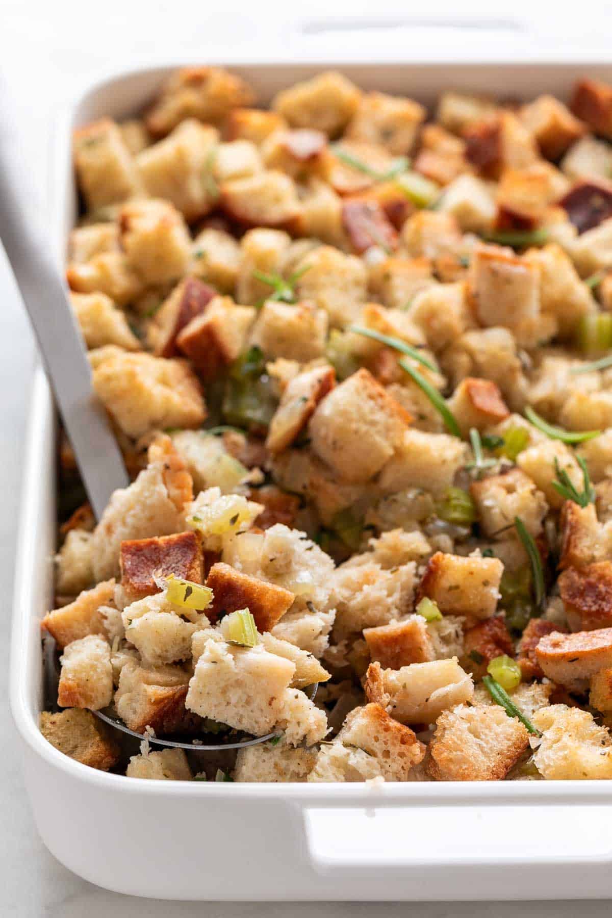 Vegan stuffing in a baking dish with a serving spoon and fresh rosemary on top.