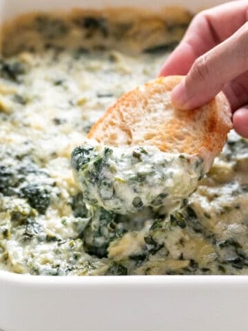 Hand dipping bread into vegan spinach artichoke dip in a baking dish.