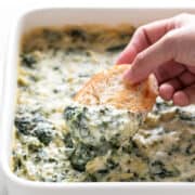 Hand dipping a slice of bread into a baking dish with vegan spinach artichoke dip.