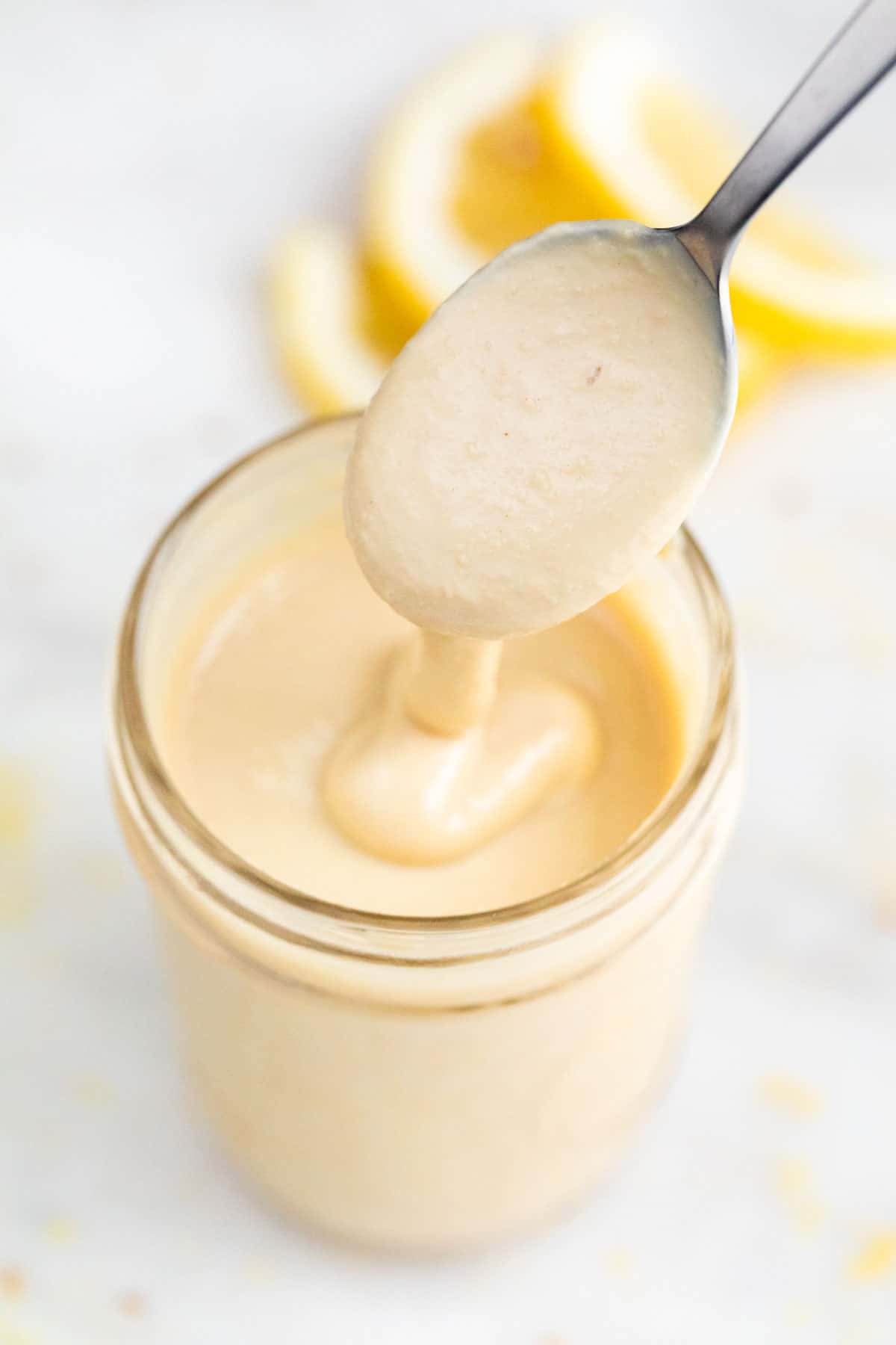 Jar of lemon tahini dressing, with lemon slices at the bottom, being spoon-drizzled.