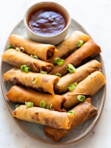 Vegan spring rolls plated with sweet and sour sauce and topped with spring onions.
