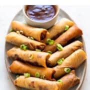 Vegan spring rolls on a plate with sweet and sour sauce garnished with spring onions.