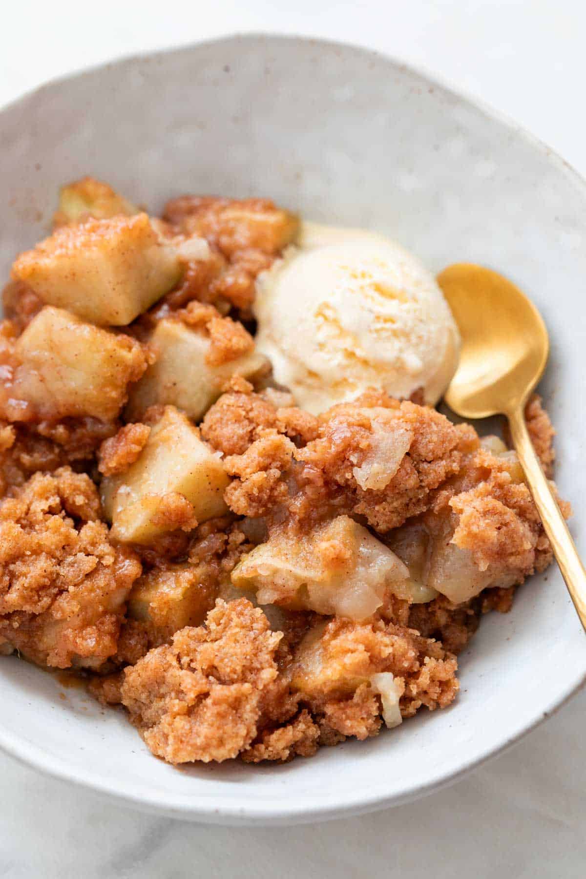 Bowl with vegan apple crumble, topped with ice cream and a spoon beside it.