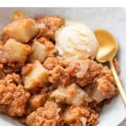 Bowl with vegan apple crumble, topped with ice cream and a spoon beside it.