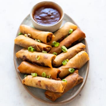 Vegan spring rolls plated with sweet and sour sauce and topped with spring onions.