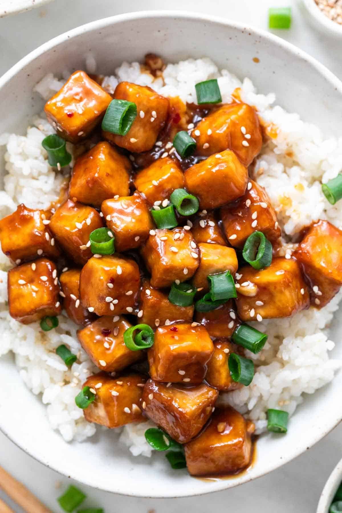 Garnished orange tofu served over rice with chopsticks on the side.