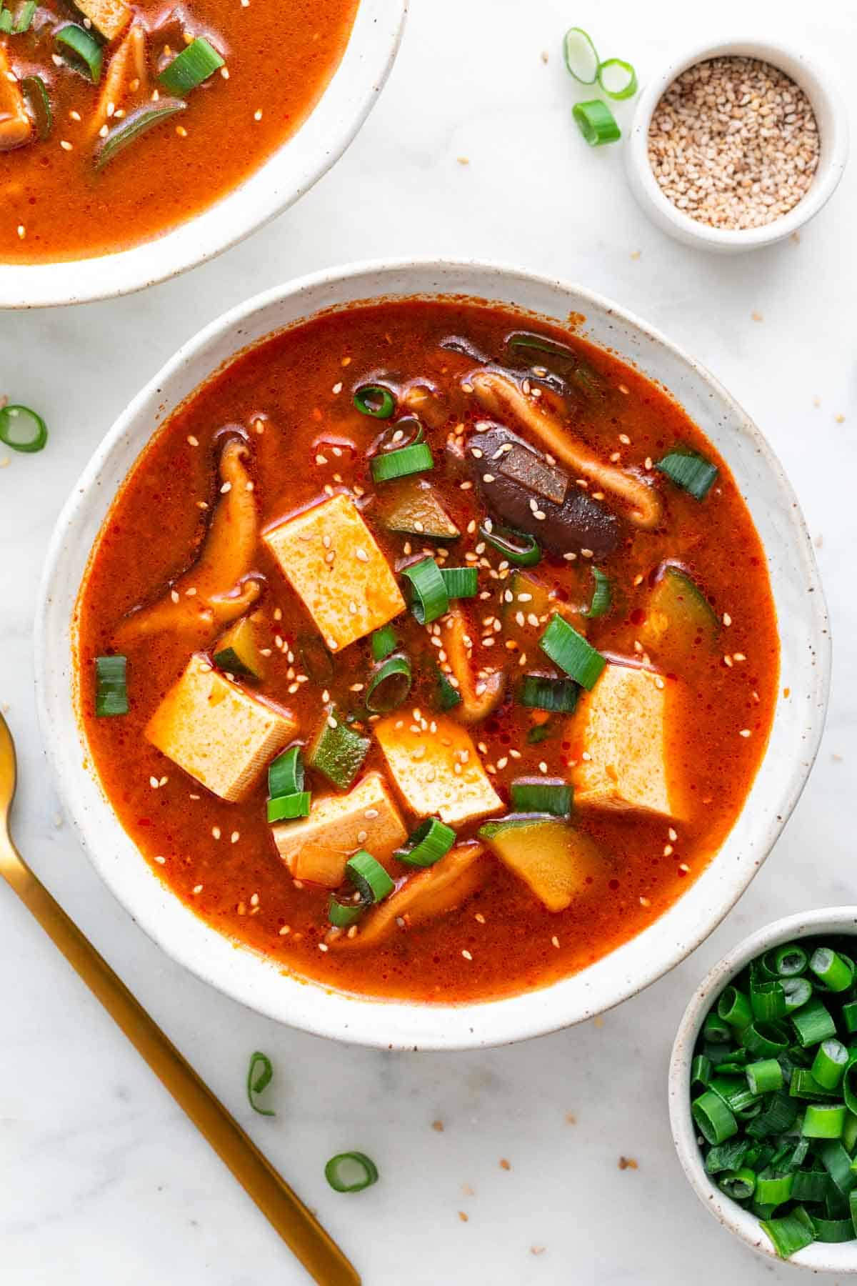 Two bowls of spicy Korean tofu soup, surrounded by additional bowls of toppings.