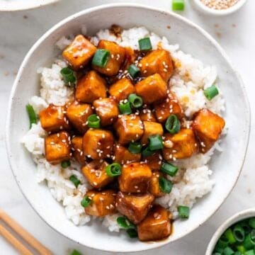 Garnished orange tofu served over rice in a bowl, with ingredients arranged around it.