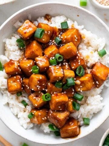 Garnished orange tofu served over rice in a bowl, with ingredients arranged around it.