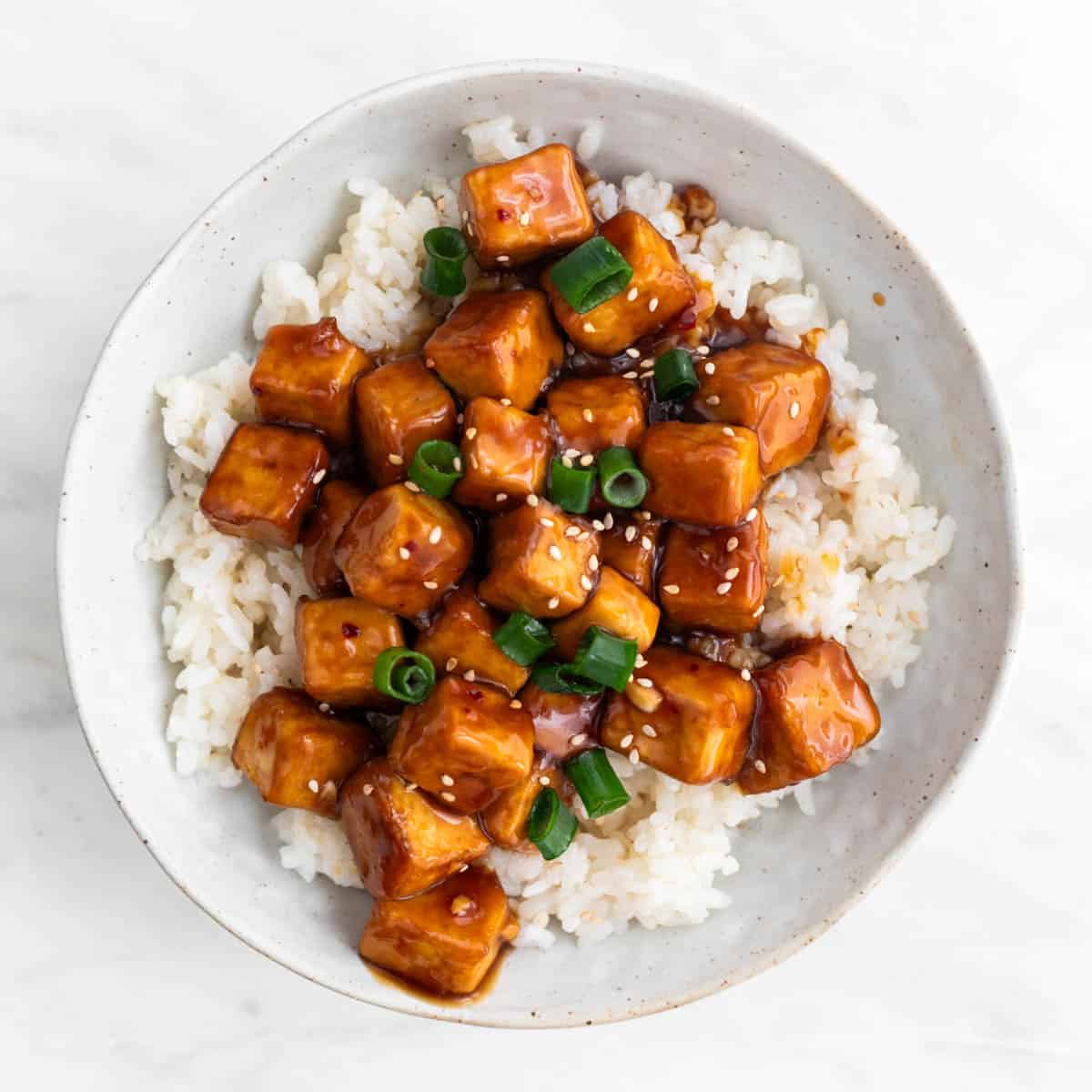 Orange tofu served over rice in a bowl, topped with sesame seeds and scallions.