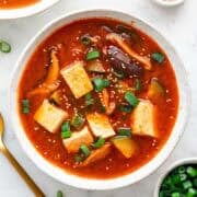 Two bowls of spicy Korean tofu soup, surrounded by bowls of toppings and a spoon.