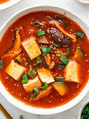 Two bowls of spicy Korean tofu soup, surrounded by bowls of toppings and a spoon.