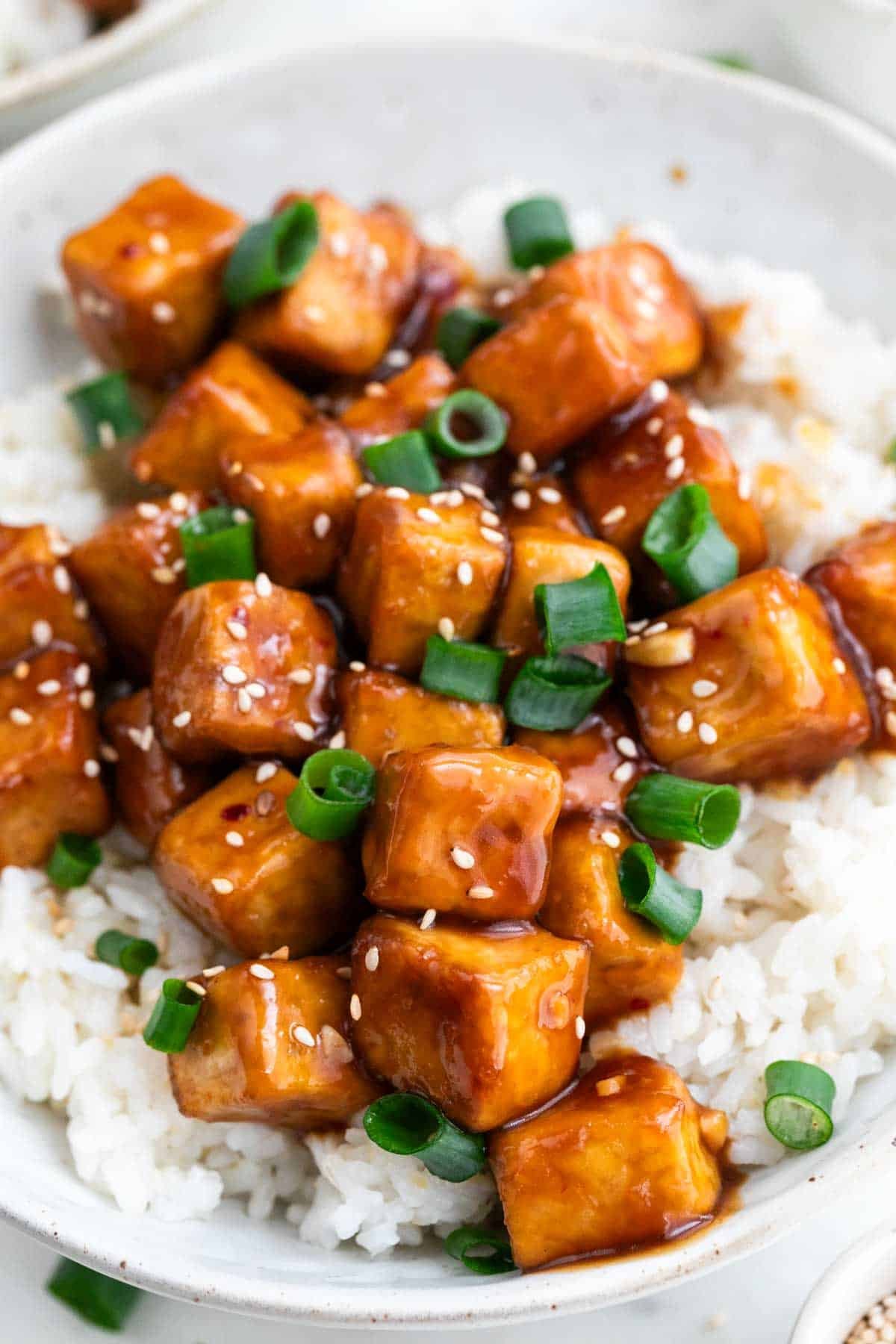 Orange tofu served in a bowl over rice, garnished with sesame seeds and scallions.