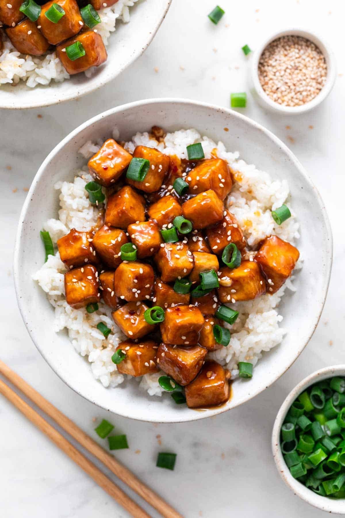 Garnished orange tofu served over rice in a bowl, with ingredients arranged around it.